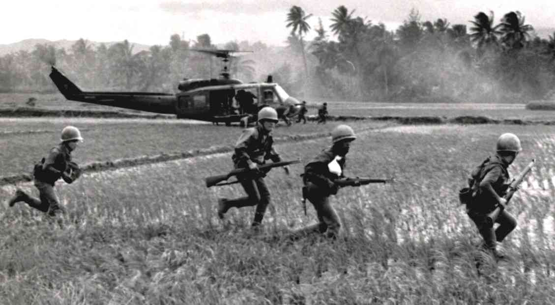 Soldiers running through a rice paddy as a helicopter lands