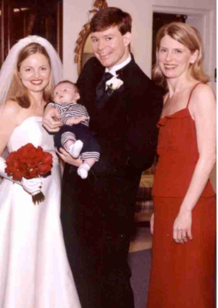 A bride and groom holding a baby beside a woman in red